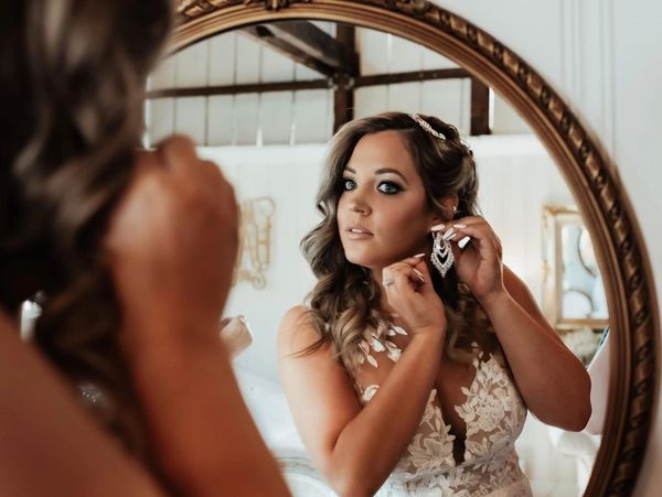 Bride adjusting earrings in front of a mirror, wearing a lace wedding dress.