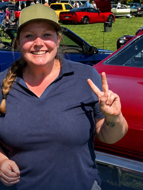 Smiling woman in cap making a peace sign at car show.