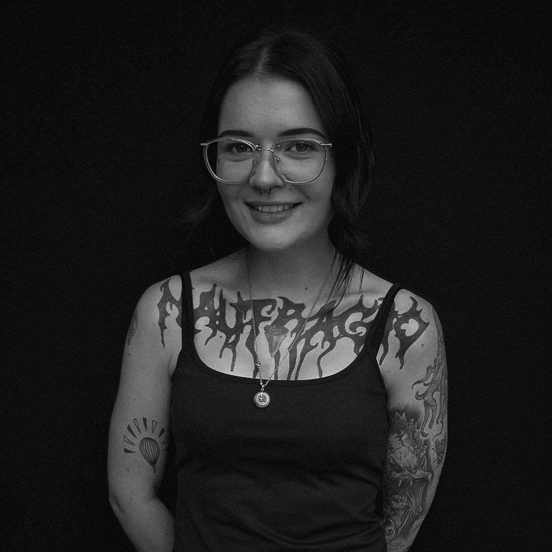 Smiling young woman with glasses and multiple tattoos, wearing a black tank top.