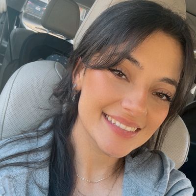 Smiling woman taking a selfie inside a car, wearing a gray jacket and brown top.