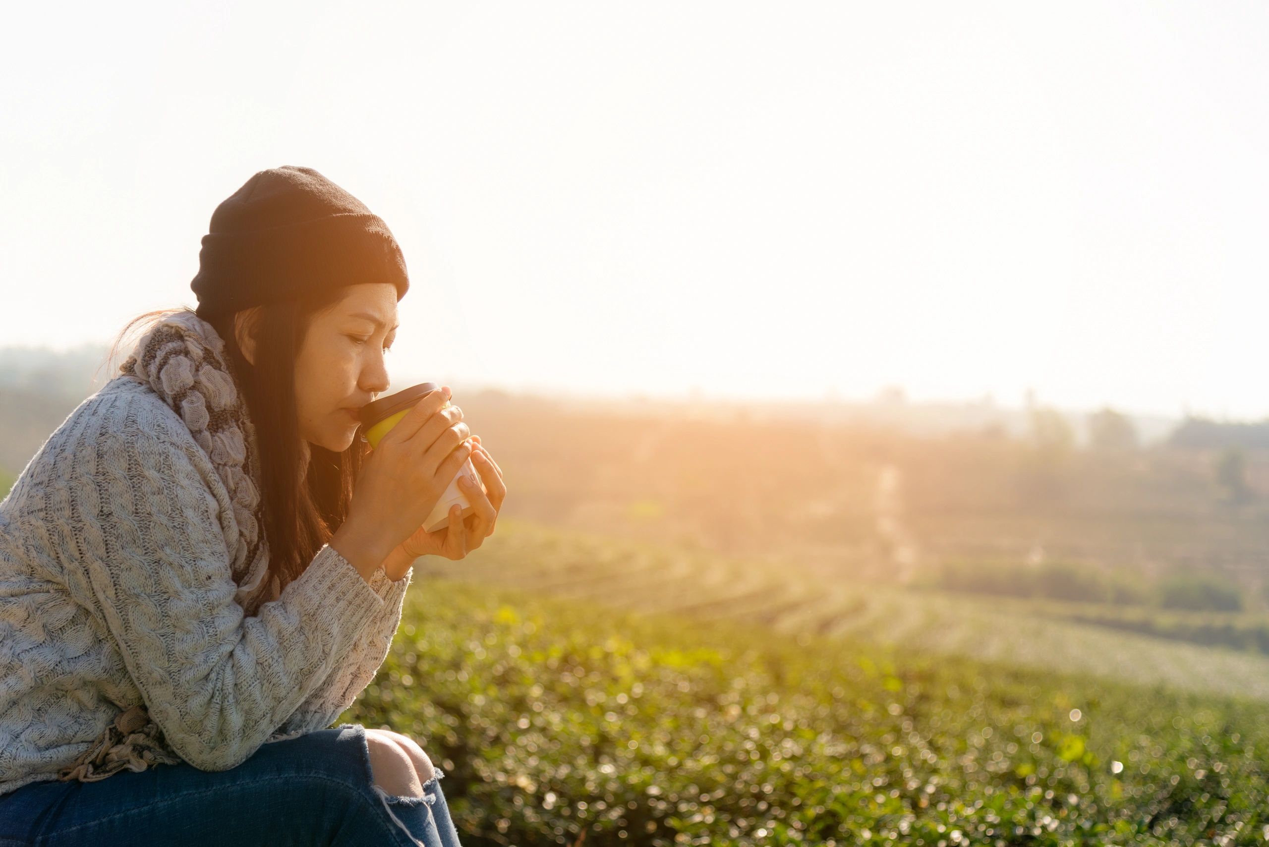 Woman in grey sweater drinking coffee/tea near open field.