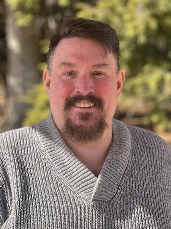 Man with a beard smiling outdoors in a gray knitted sweater.
