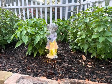 Garden statue of a girl pouring water surrounded by green plants and mulch.