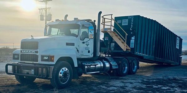 White Mack truck hauling a large green container at sunset on a dirt road.