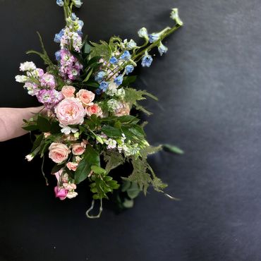 A hand holding a delicate bouquet of pink and blue flowers with greenery against a dark background.