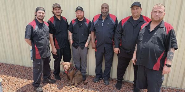 Six men in matching work uniforms with a dog pose outside a building.