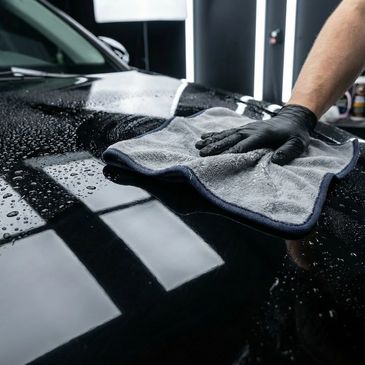 Person wearing gloves polishing a wet black car with a microfiber cloth.