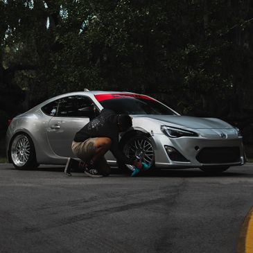 A person cleaning the front wheel of a silver sports car on a road.