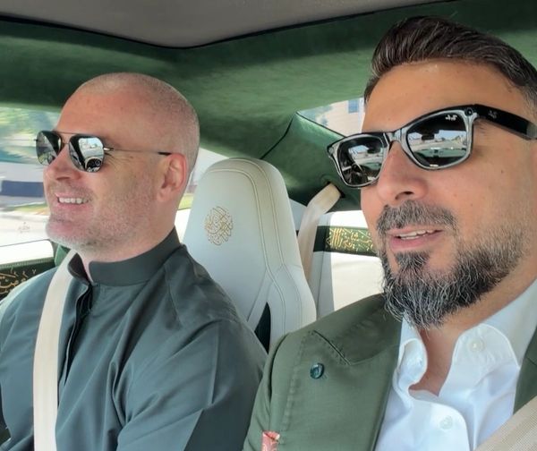 Two men wearing sunglasses sitting inside a car, smiling.