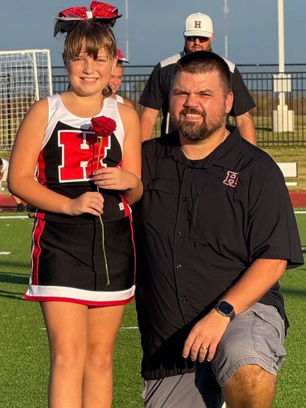 A young cheerleader and a man kneeling on a sports field smile for the photo.