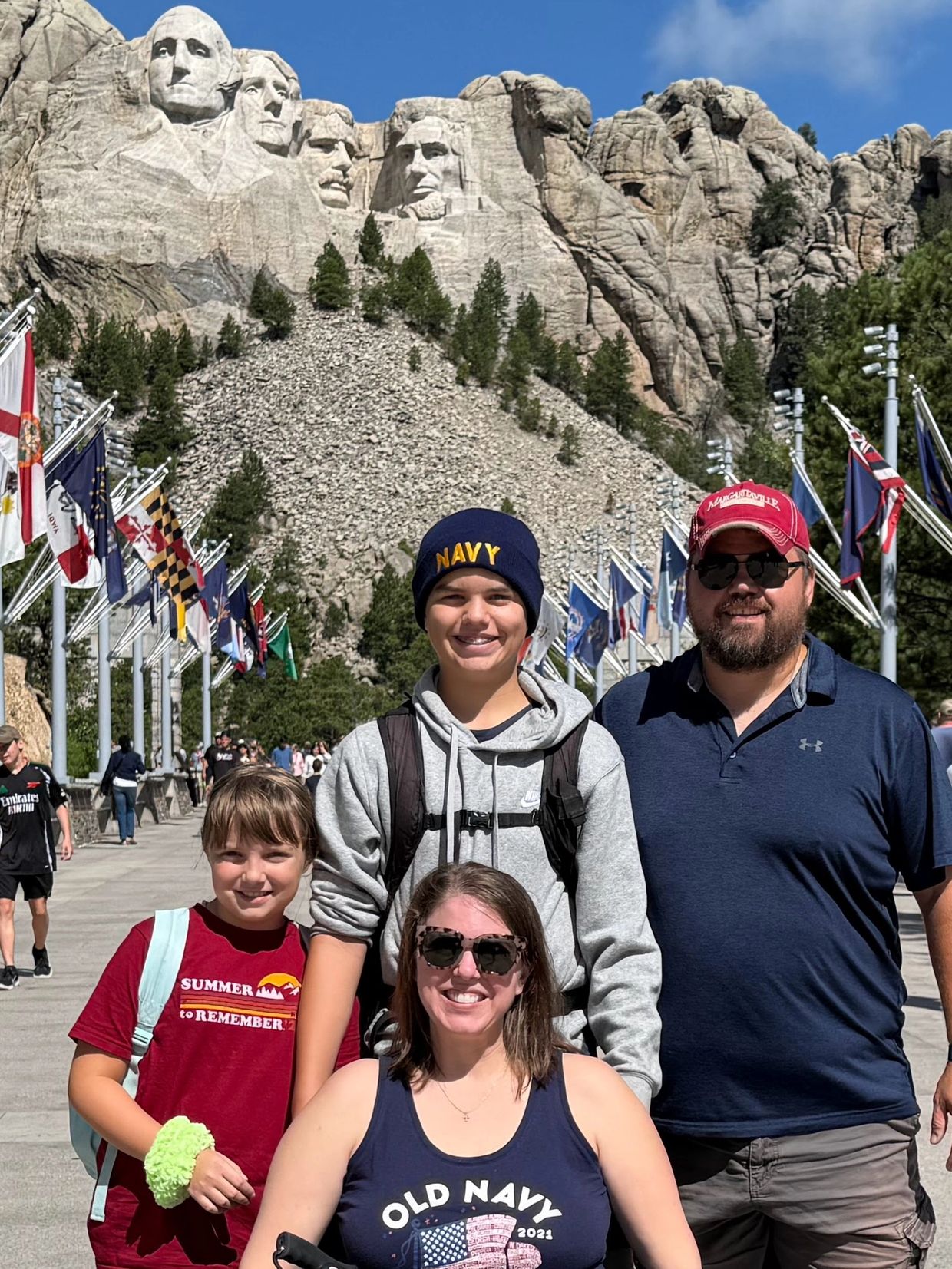 Family poses happily in front of Mount Rushmore with flags lining the walkway.