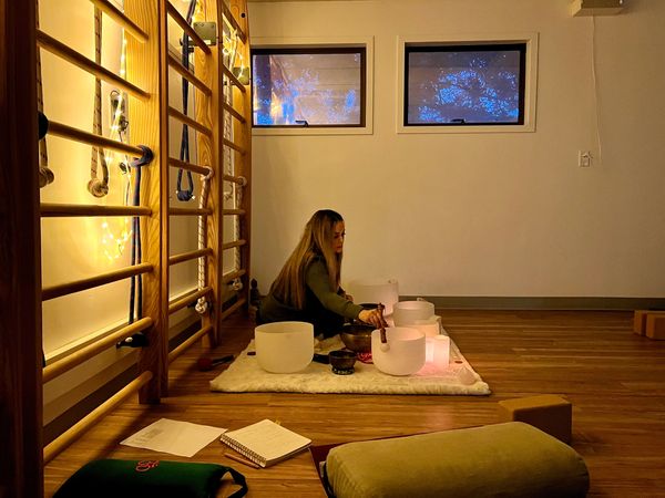 A woman practices sound healing with crystal bowls in a cozy room with warm lighting.
