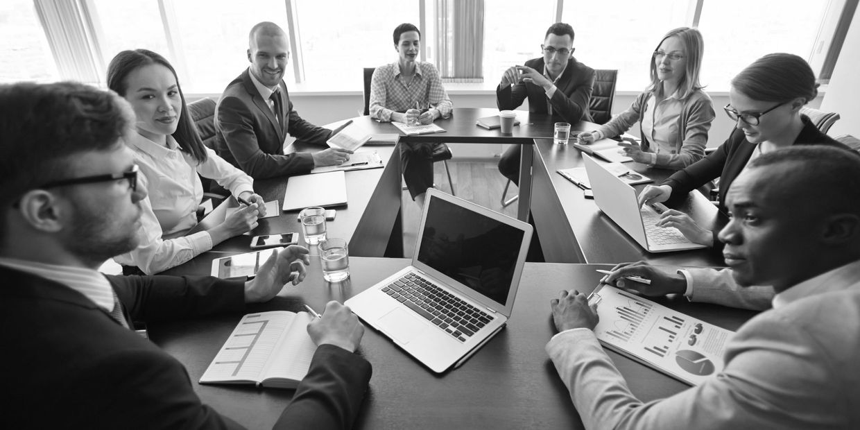 A diverse group of professionals in a business meeting around a table with laptops and documents.