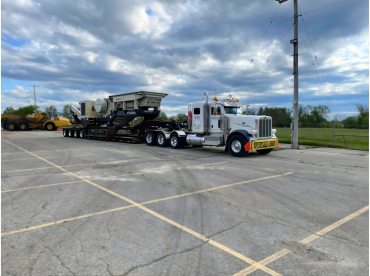 A white semi-truck parked in an empty lot carrying heavy machinery on a flatbed trailer.