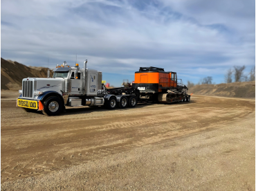 White semi-truck hauling an orange construction vehicle on a dirt road.