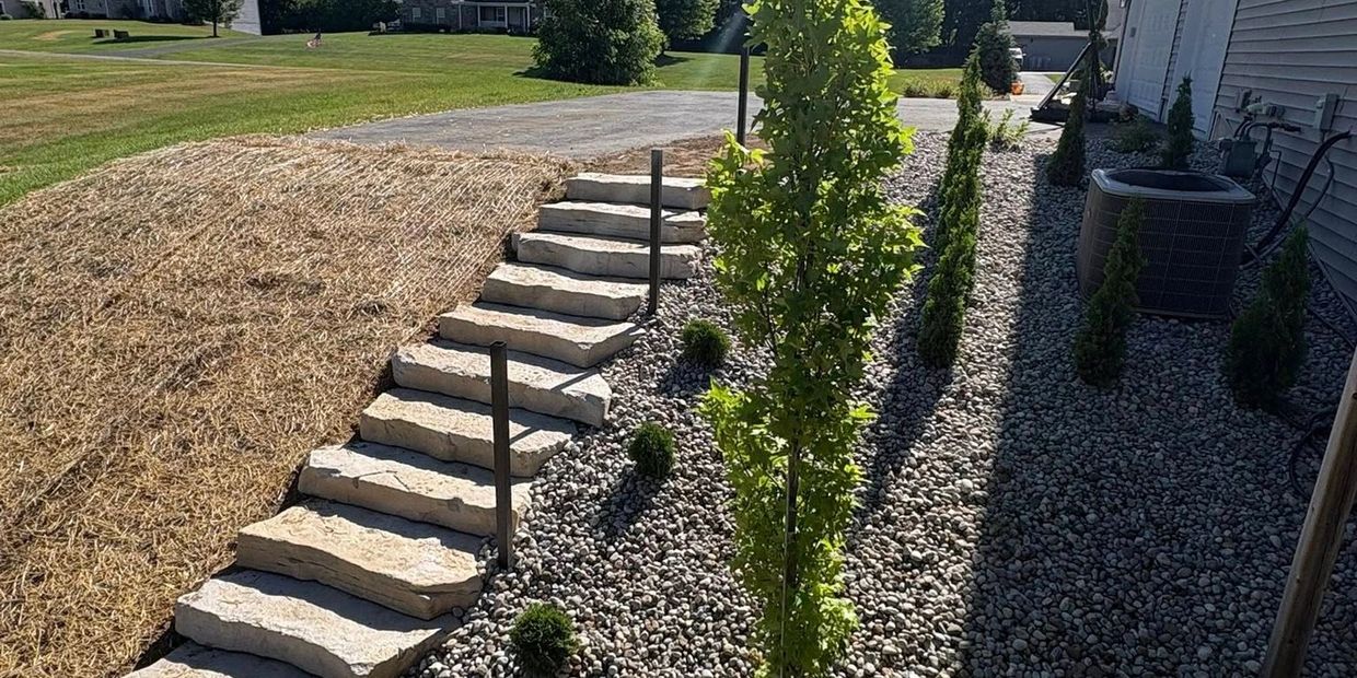 Stone steps with metal railings lead up from a landscaped area with rocks and small trees beside a house.