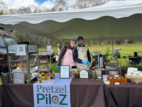 Two vendors at Pretzel Pilz food stall with pretzels and condiments.