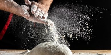 Hands dusting flour over dough on a wooden surface with a black background.