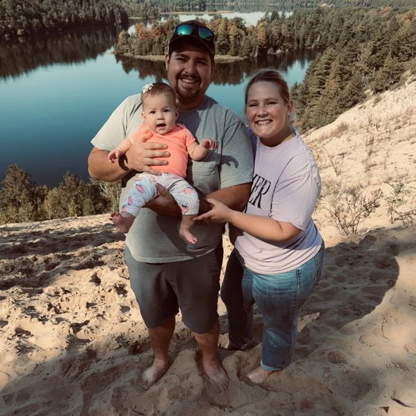A happy family of three posing barefoot on a sandy hill by a lake.