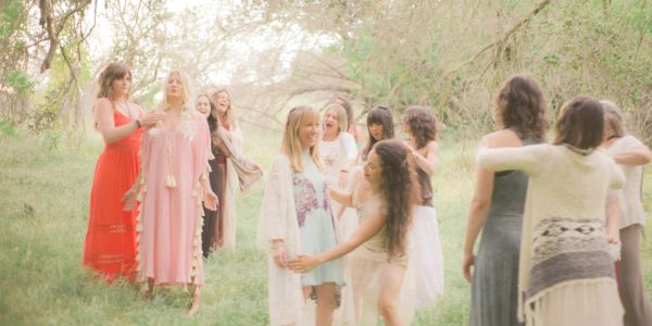 Group of women in flowing dresses enjoying an outdoor gathering in a sunlit forest.