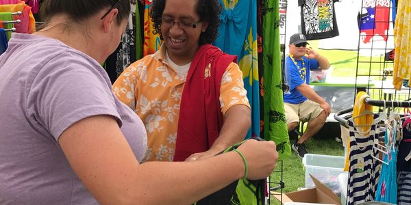 Two people smiling and shopping for colorful clothes at an outdoor market stall.
