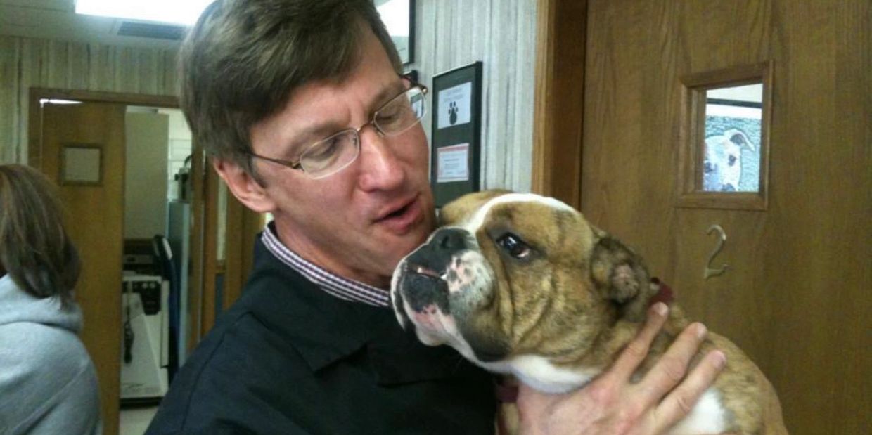 Man lovingly holding a bulldog inside a room with another dog peeking through a door window.