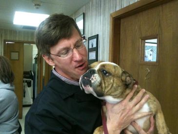 Man lovingly holding a bulldog inside a room with another dog peeking through a door window.