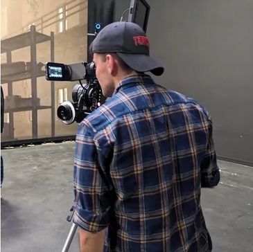 Man in plaid shirt filming with professional camera indoors.