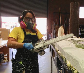 Person wearing protective gear using a power tool on a foam block indoors.