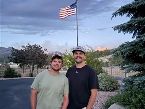 Two men smiling outdoors with an American flag and hills in the background.