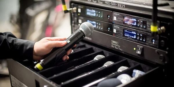 Hand holding a microphone near wireless audio equipment with multiple microphones stored.