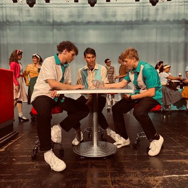 Three young men in retro outfits sit at a diner table on stage.