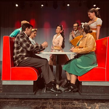 Actors in vintage diner costumes gathered around a red booth on stage.
