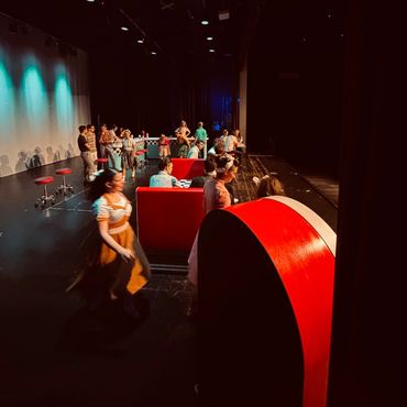 Actors rehearsing a scene on a stage with red furniture and dim lighting.
