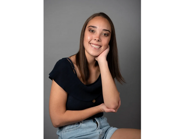 Young woman smiling, resting her head on her hand, wearing a black top and denim shorts.