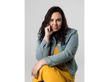 Woman with curly hair wearing a yellow dress and denim jacket, posing with a confident expression.