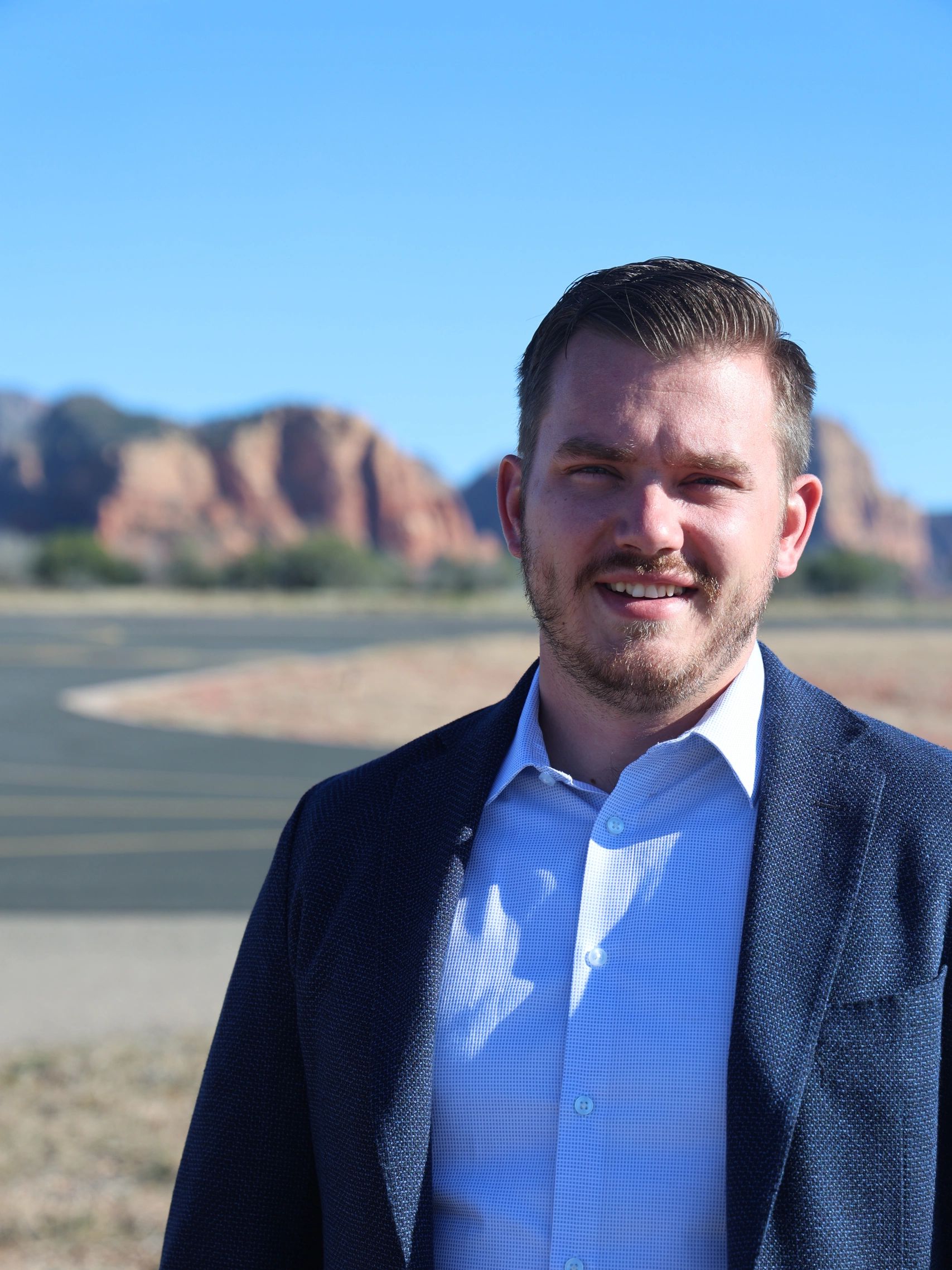 Man in blue blazer smiling outdoors with mountains in the background.