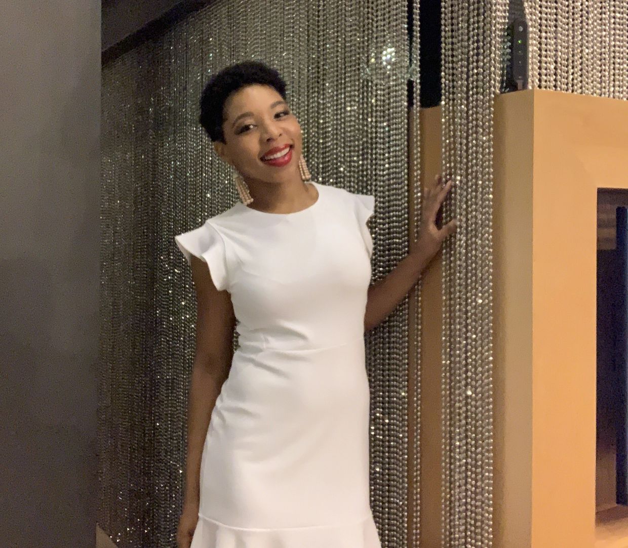 Woman in a white dress smiling by a sparkling beaded curtain.