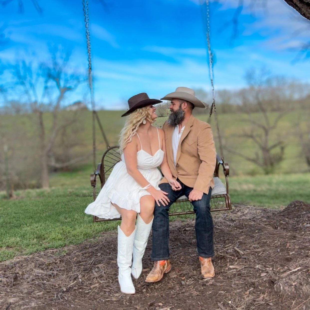 Couple in cowboy hats sitting on a swing under a large tree in a scenic outdoor setting.