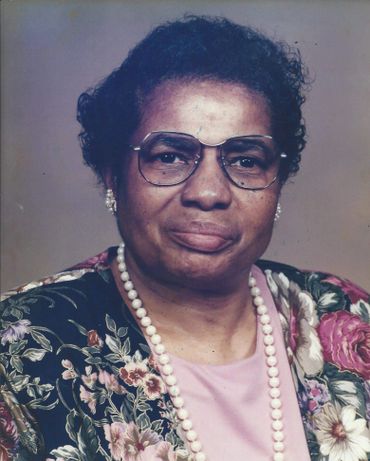 Portrait of an elderly woman wearing glasses and floral attire with a pearl necklace.