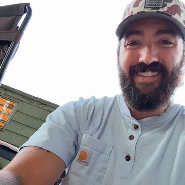 Bearded man smiling outdoors wearing a camo hat and light blue shirt.