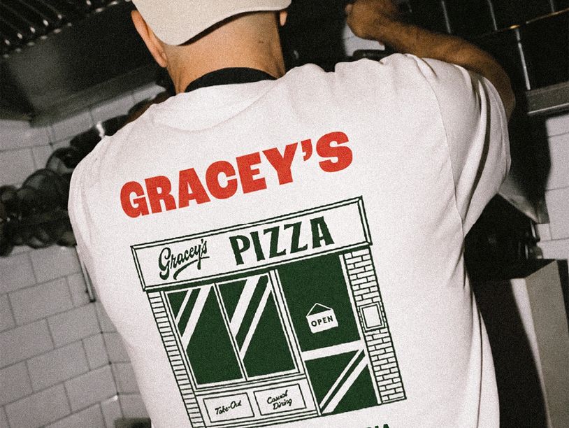 A person wearing a Gracey's Pizza shirt and hat in a kitchen.