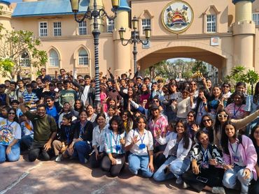 Large group posing happily in front of a castle-like building on a sunny day.