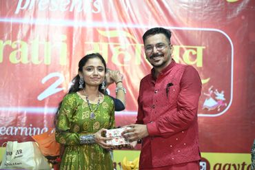 A man in a red kurta presents a gift to a woman in a green dress at an event.