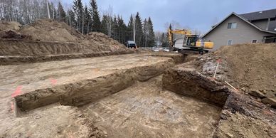 Excavator digging a large foundation pit near a house on a cloudy day.