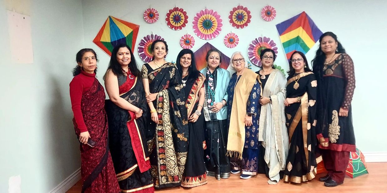 A group of women dressed in traditional sarees posing indoors with colorful decorations on the wall.