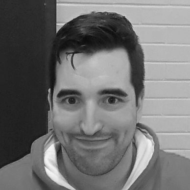 A man smiling with a hoodie against a brick wall background.
