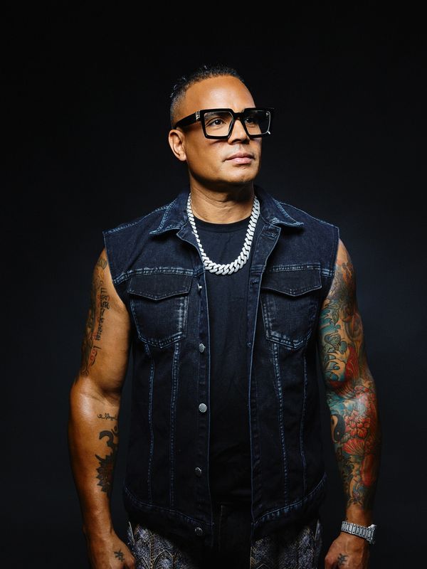 Man with large glasses, tattoos, and a silver chain wearing a sleeveless denim jacket.