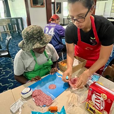 Two people preparing food with salami and snacks on a table indoors.