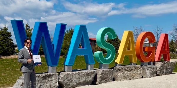 Man in suit stands beside colorful WASAGA sign outdoors on a sunny day.